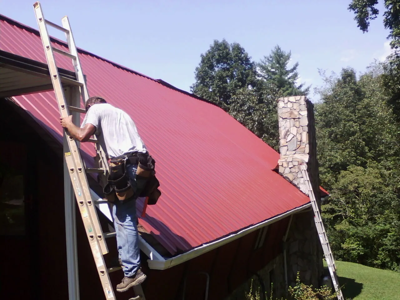 Roofer on a ladder beside a red metal roof and stone chimney — Soffit & Fascia Repair by Roofing Brothers in Charlottesville